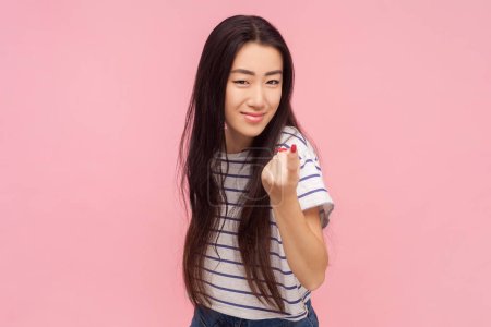 Portrait of playful woman with long brunette hair showing beckoning gesture saying come here, wearing striped T-shirt. Indoor studio shot isolated on pink background.