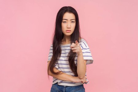 Porträt einer wütenden Frau mit langen brünetten Haaren, die mit erhobenem Zeigefinger und gestreiftem T-Shirt dasteht. Indoor-Studio isoliert auf rosa Hintergrund aufgenommen.