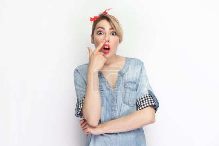 Portrait of funny crazy blonde woman wearing blue denim shirt and red headband standing keeps finger in nose, looking with foolish expression. Indoor studio shot isolated on gray background.