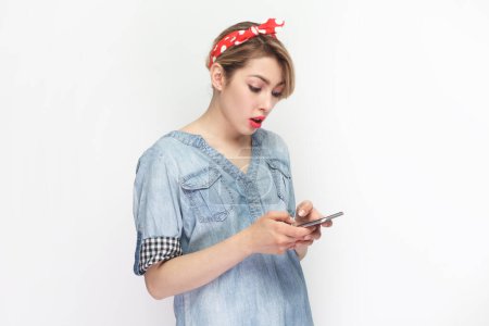 Téléchargez les photos : Portrait latéral d'une femme blonde sérieuse et concentrée portant une chemise en denim bleu et un bandeau rouge debout à l'aide d'un téléphone portable, vérifiant les e-mails. Studio intérieur tourné isolé sur fond gris. - en image libre de droit
