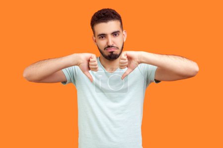 Photo for Portrait of displeased sad unhappy young bearded man wearing T-shirt standing showing thumbs down looking at camera with negative feelings. Indoor studio shot isolated on orange background. - Royalty Free Image