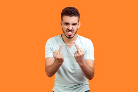 Portrait of strict impolite young bearded man wearing T-shirt showing middle fingers fuck off gestures arguing with somebody. Indoor studio shot isolated on orange background.