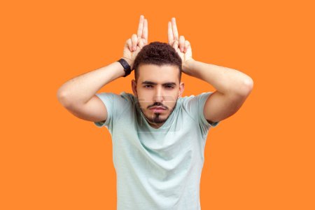 Photo for Portrait of angry young bearded man wearing T-shirt showing devil horns demonstrating arguing with somebody hate expression. Indoor studio shot isolated on orange background. - Royalty Free Image
