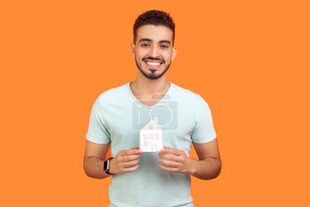 Téléchargez les photos : Portrait d'un jeune homme barbu souriant portant un T-shirt debout montrant une maison en papier rêvant de son propre appartement. Studio intérieur tourné isolé sur fond orange. - en image libre de droit