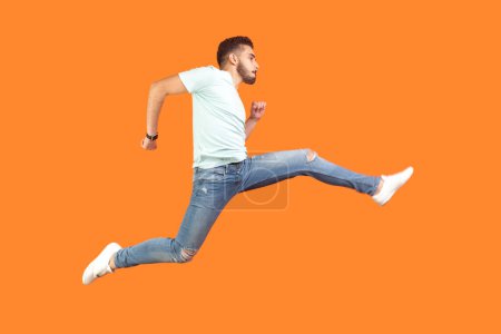 Photo for Full length portrait of funny energetic carefree young bearded man wearing T-shirt jumping running in air being hurry. Indoor studio shot isolated on orange background. - Royalty Free Image