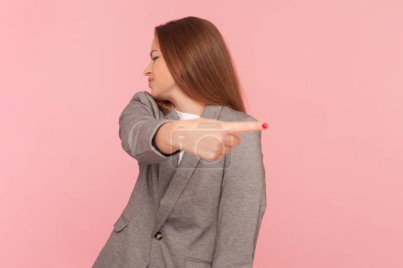 Photo for Portrait of offended upset woman with brown hair turning her face and pointing away, saying go away, showing direction, wearing business suit. Indoor studio shot isolated on pink background. - Royalty Free Image