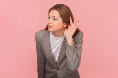 Photo for Portrait of attractive woman with brown hair keeps hand near ear and listening gossips, having hearing problems, wearing business suit. Indoor studio shot isolated on pink background. - Royalty Free Image