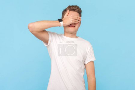 Téléchargez les photos : Portrait de l'homme effrayé portant un t-shirt blanc debout couvrant les yeux de paume, voit quelque chose d'effrayant ou honteux, a la phobie. Studio intérieur tourné isolé sur fond bleu - en image libre de droit