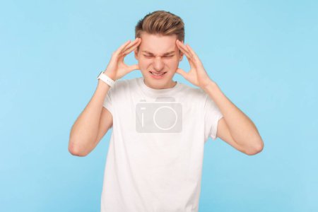 Photo for Portrait of sick unhealthy ill man wearing white t-shirt massaging painful temples, suffering terrible headache, frowning face. Indoor studio shot isolated on blue background - Royalty Free Image