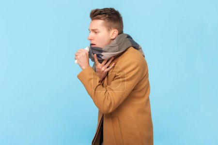 Téléchargez les photos : Portrait d'un jeune homme malade portant une veste chaude blanche et un foulard toussant debout, présentant des symptômes de grippe, une grippe, une température élevée. Studio intérieur tourné isolé sur fond bleu - en image libre de droit