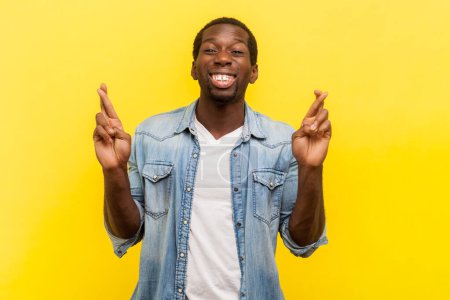 Photo for Portrait of smiling happy man standing with crossed fingers making wish praying for good luck waiting for success, wearing denim casual shirt. Indoor studio shot isolated on yellow background. - Royalty Free Image
