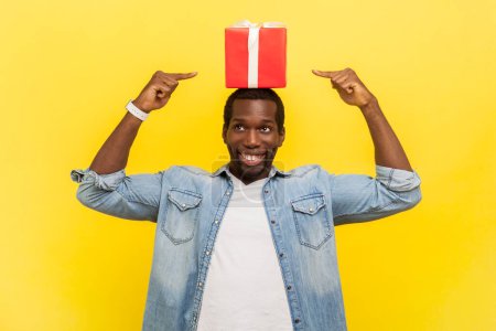 Photo for Portrait of satisfied positive optimistic man standing with wrapped present box on head and pointing at gift smiling, wearing denim casual shirt. Indoor studio shot isolated on yellow background. - Royalty Free Image