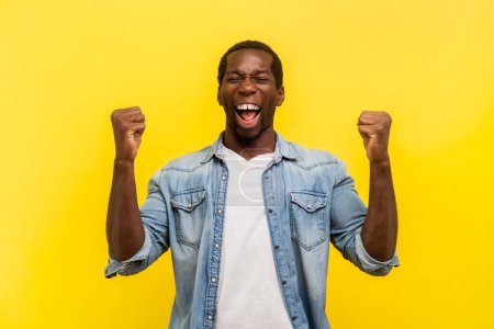 Photo for Portrait of overjoyed happy delighted man clenched fists screaming with happiness and amazement celebrating success, wearing denim casual shirt. Indoor studio shot isolated on yellow background. - Royalty Free Image