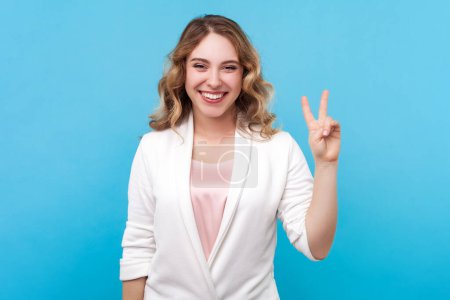 Photo for Portrait of charming positive blond woman with wavy hair standing showing v sign victory peace gesture looking with glad expression, wearing white shirt. Indoor studio shot isolated on blue background - Royalty Free Image