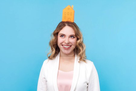Photo for Portrait of happy blond woman with wavy hair standing with paper house on head dreaming about her own apartment, real estate, wearing white shirt. Indoor studio shot isolated on blue background. - Royalty Free Image