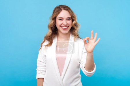 Photo for Portrait of cute positive smiling blond woman with wavy hair looking at camera showing okay gesture ok sign, wearing white shirt. Indoor studio shot isolated on blue background. - Royalty Free Image