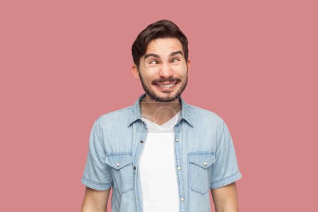 Portrait of hilarious crazy bearded man in casual style shirt standing crosses eyes, makes funny grimace, pretends to be little fool, being childish. Indoor studio shot isolated on pink background.