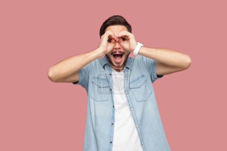 Photo for Surprised bearded man standing looking through binoculars hand gesture, exploring discovering something unbelievable, expressing amazement. Indoor studio shot isolated on pink background. - Royalty Free Image