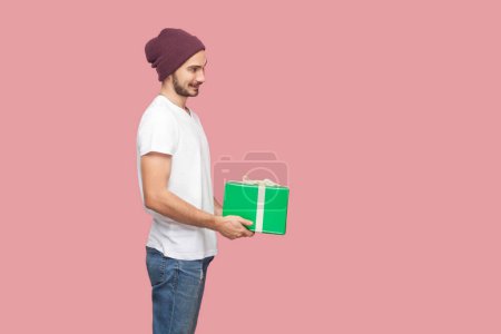 Photo for Portrait of happy positive satisfied bearded man in white T-shirt and beany hat standing standing with green wrapped present box, congratulating. Indoor studio shot isolated on pink background. - Royalty Free Image