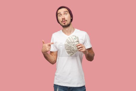 Photo for Portrait of astonished surprised bearded man in white T-shirt and beany hat standing pointing at dollar banknotes, looking at camera with big eyes. Indoor studio shot isolated on pink background. - Royalty Free Image