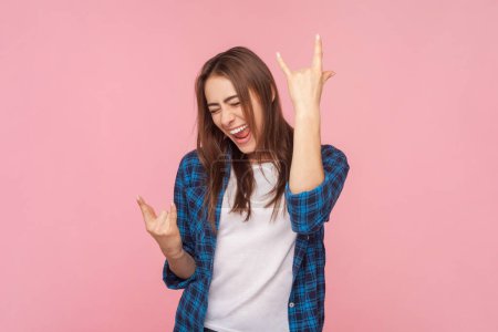 Photo for Portrait of extremely excited brown haired woman screaming with happy emotions on concert showingrock and roll gesture. Indoor studio shot isolated on pink background. - Royalty Free Image