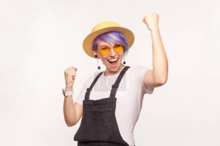 Téléchargez les photos : Portrait d'une femme extrêmement heureuse aux cheveux violets avec lunettes de soleil et poings serrés au chapeau criant de bonheur célébrant son succès. Studio intérieur tourné isolé sur fond blanc. - en image libre de droit
