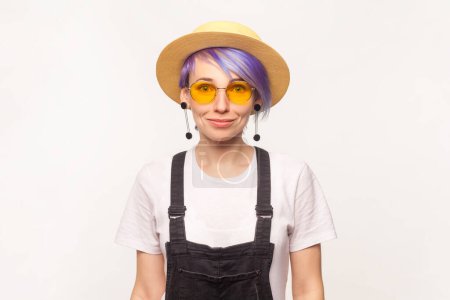 Photo for Portrait of smiling positive hipster woman with violet hair in sunglasses and hat looking at came with pleased emotion. Indoor studio shot isolated on white background. - Royalty Free Image