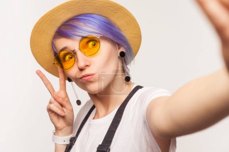 Foto de Retrato de la blogger divertida mujer cool con pelo violeta en gafas de sol y sombrero haciendo selfie POV mostrando v signo de mueca. Estudio interior plano aislado sobre fondo blanco. - Imagen libre de derechos