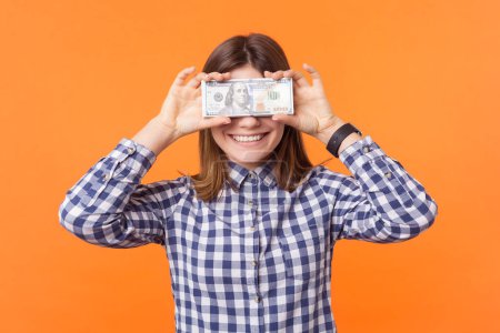 Photo for Portrait of happy shy rich woman with brown hair covering her eyes with dollar banknotes smiling to camera, wearing checkered shirt. Indoor studio shot isolated on orange background - Royalty Free Image