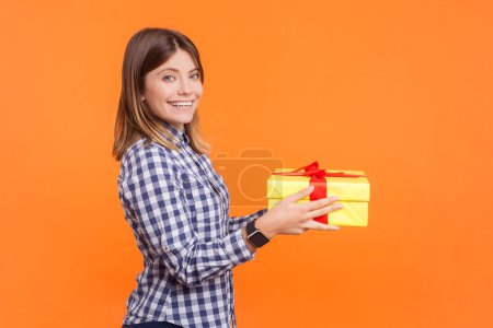 Téléchargez les photos : Portrait de profil de souriante femme heureuse et optimiste aux cheveux bruns offrant un cadeau au petit ami regardant la caméra, portant une chemise à carreaux. Studio intérieur tourné isolé sur fond orange - en image libre de droit