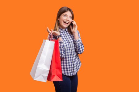 Portrait of cute charming woman with brown hair holding shopping bags and talking phone, boasting purchases, wearing checkered shirt. Indoor studio shot isolated on orange background