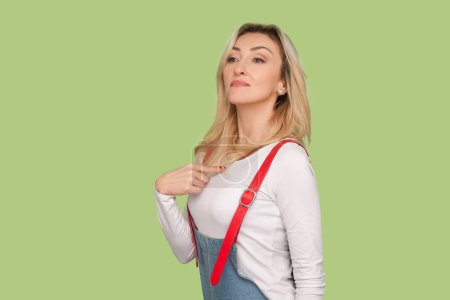 Photo for Portrait of proud selfish egoistic adult blond woman standing pointing at herself, looking at camera with arrogant face, wearing denim overalls. Indoor studio shot isolated on light green background - Royalty Free Image