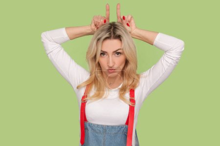 Photo for Portrait of angry aggressive adult blond woman making devil cow horns, being ready to attack, arguing with somebody, wearing denim overalls. Indoor studio shot isolated on light green background - Royalty Free Image
