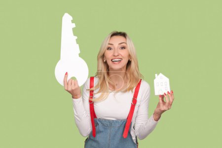 Portrait of satisfied cute adult blond woman holding paper key and house in hands, looking at camera, relocate, wearing denim overalls. Indoor studio shot isolated on light green background