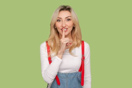 Photo for Portrait of cute charming adult blond woman keeps finger near mouth, keeping secret, looking at camera, wearing denim overalls. Indoor studio shot isolated on light green background - Royalty Free Image