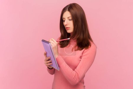 Photo for Clever serious woman with brown hair writing in notebook, completing to-do list or studying, having concentrated expression, wearing rose turtleneck. Indoor studio shot isolated on pink background - Royalty Free Image
