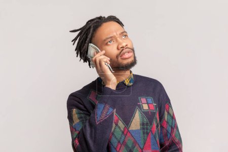 Photo for Portrait of funny positive playful african-american man with dreadlocks and beard standing with dollar banknotes near ear, pretending talking on phone. Indoor studio shot isolated on gray background. - Royalty Free Image