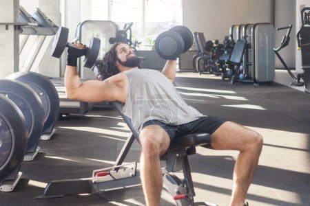 Photo for Portrait of strong hard working man with long curly hair working out in gym, doing exercising on bench and holding dumbbells with raised arm, training biceps. Indoor shot. - Royalty Free Image