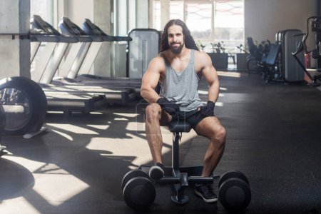 Photo for Portrait of strong young adult man with long curly hair athlete working out in fitness club sitting on bench dong exercises for his body, looking at camera with confident expression. Indoor shot. - Royalty Free Image