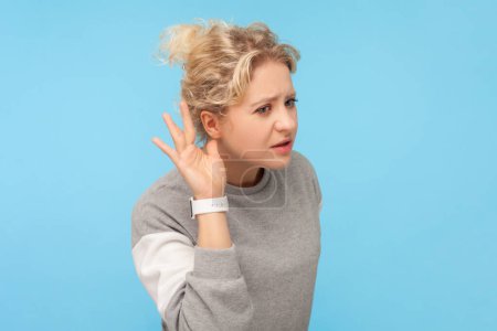 Photo for Portrait of serious blonde woman holding hand near her ear, trying to hear silent talking, private conversation, wearing gray sweatshirt. Indoor studio shot isolated on blue background. - Royalty Free Image