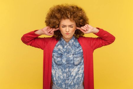 Stop making this terrible noise. Stressed nervous woman with Afro hairstyle closing eyes and plugging ears with fingers, annoyed with loud sounds. Indoor studio shot isolated on yellow background.