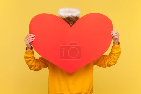 Photo for Portrait of angelic anonymous woman with nimb above head with Afro hairstyle hiding behind big red heart, wearing casual style hoodie. Indoor studio shot isolated on yellow background. - Royalty Free Image