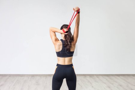 Back view of young sporty woman wearing black tank top and leggings, doing exercise for triceps with latex resistance band. Full length indoor shot against white wall.