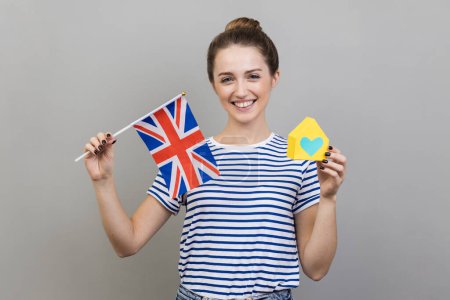 Photo for Portrait of self confident smiling woman wearing striped T-shirt holding Great Britain flag and paper house, dreaming to buy accommodation in England. Indoor studio shot isolated on gray background. - Royalty Free Image