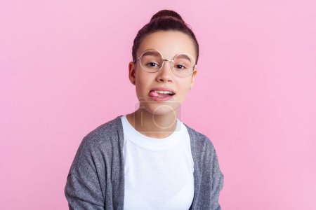 Retrato de adolescente divertida con peinado moño en ropa casual y gafas de pie mirando a la cámara que muestra la lengua hacia fuera. Estudio interior plano aislado sobre fondo rosa.