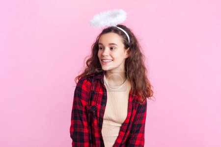 Portrait of charming teenage girl with wavy hair in red checkered shirt and nimb over head looking away with dreamy expression. Indoor studio shot isolated on pink background.