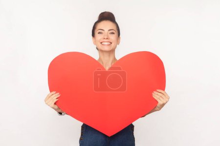 Photo for Portrait of romantic cheerful woman with hair bun holding big red heart, looking at camera with toothy smile, expressing love, wearing denim overalls. Indoor studio shot isolated on white background - Royalty Free Image