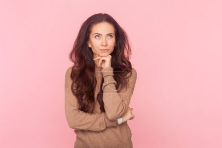 Photo for Portrait of pensive woman with wavy hair, holding her chin thinking about future, looking away, wearing wearing brown pullover. Indoor studio shot isolated on pink background - Royalty Free Image