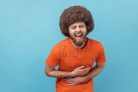 Porträt eines lachenden Mannes mit Afro-Frisur in orangefarbenem T-Shirt, den Bauch haltend und in wahnsinnig hysterischem Lachen gekrümmt, aufrichtige freudige Emotionen. Indoor Studio aufgenommen isoliert auf blauem Hintergrund.