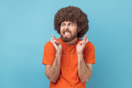 Portrait of man with Afro hairstyle in orange T-shirt standing with crossed fingers for good luck, heartily making wish, praying and hoping for miracle. Indoor studio shot isolated on blue background.